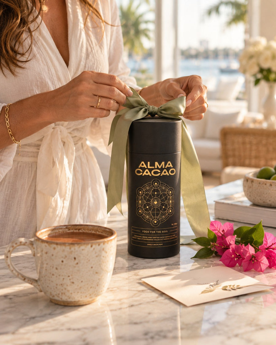 Hands tying a ribbon around a canister of Alma Cacao as a gift, with a cup of ceremonial cacao and bougainvillea in Palm Beach