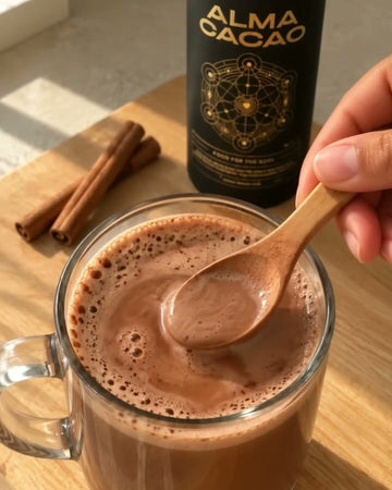 A clear glass mug of chai spiced cacao with thick foam, a wooden spoon, cinnamon sticks and the black Alma Cacao canister behind it