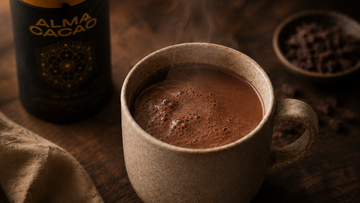 A steaming ceramic mug of thick, opaque ceremonial cacao beside an Alma Cacao canister on a warm wooden table