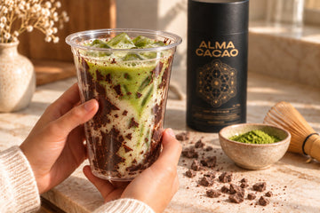 Hands holding a clear cup lined with a cracked cacao shell filled with iced matcha latte, beside the Alma Cacao canister, a bowl of matcha and a bamboo whisk