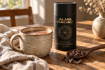 A stoneware mug of hot cacao beside the Alma Cacao canister on a wooden table in early morning light.